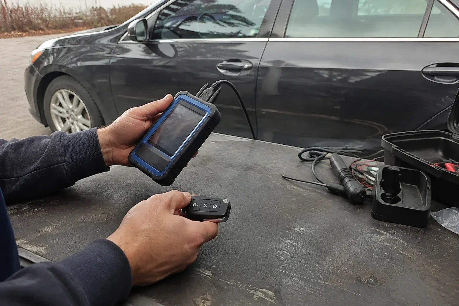 Hands holding a blue-edged electronic device and car-key fob at a worktable beside a parked car. — Transponder Key Programming, Aurora